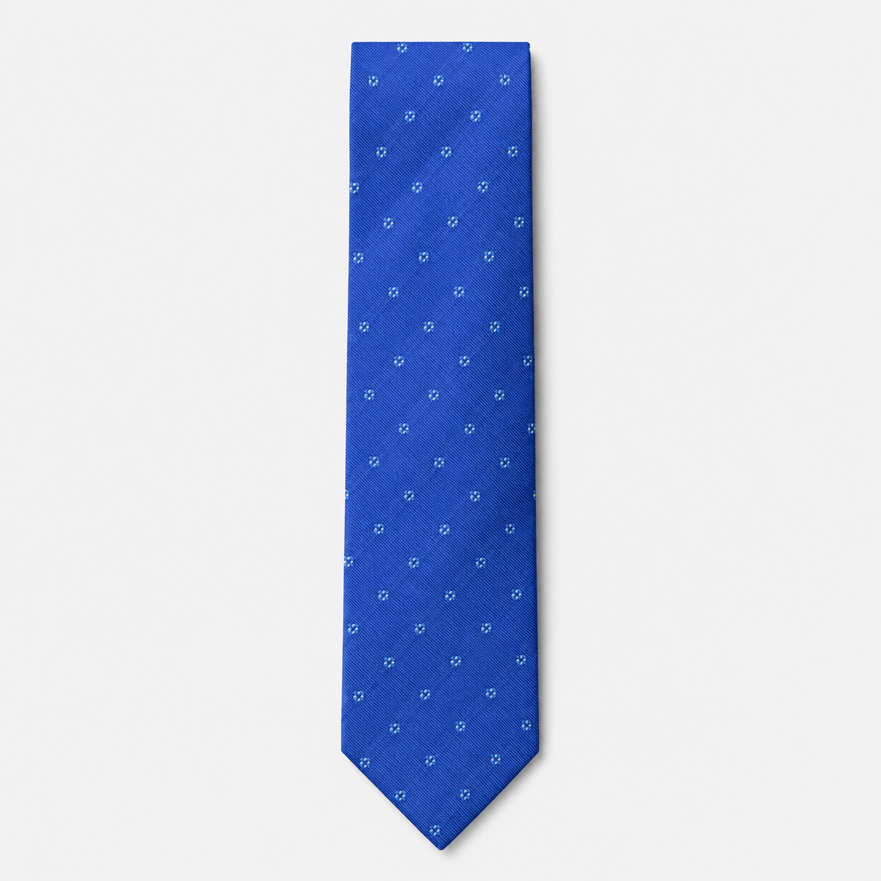 The Bellagio Tie, a royal blue silk tie with small light blue and white floral-dot motifs on a light grey background.